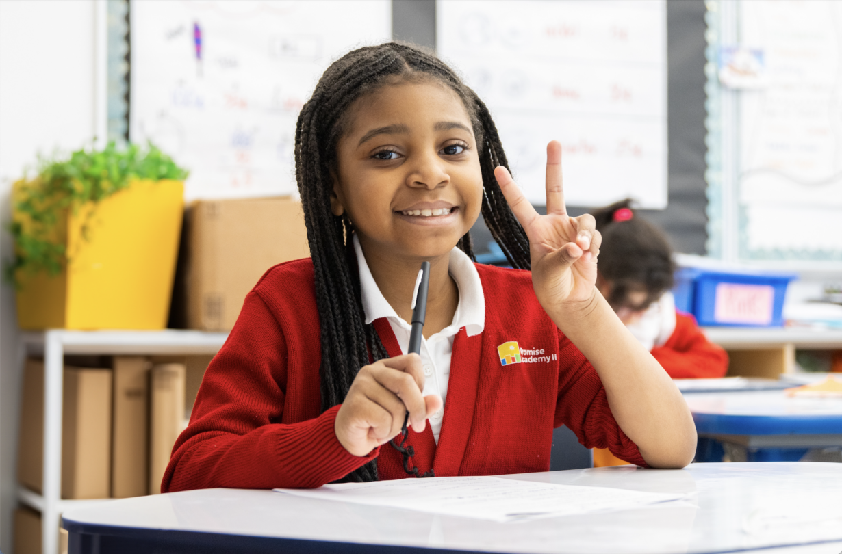 Girl wearing red cardigan making a peace sign with left hand and sitting at desk in classroom.